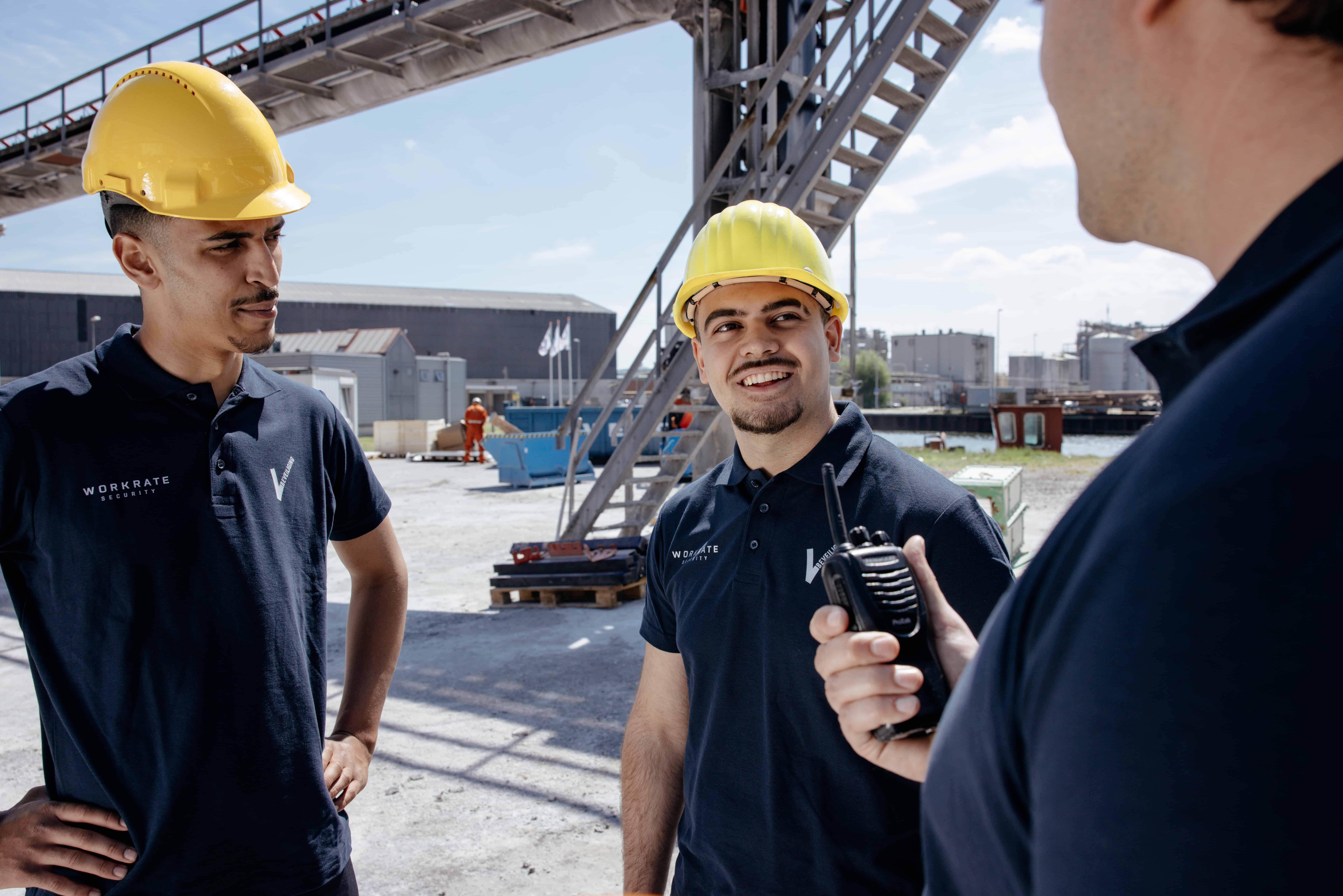Drie beveiligers met gele helmen hebben een gesprek terwijl één van hen een portofoon vasthoudt.