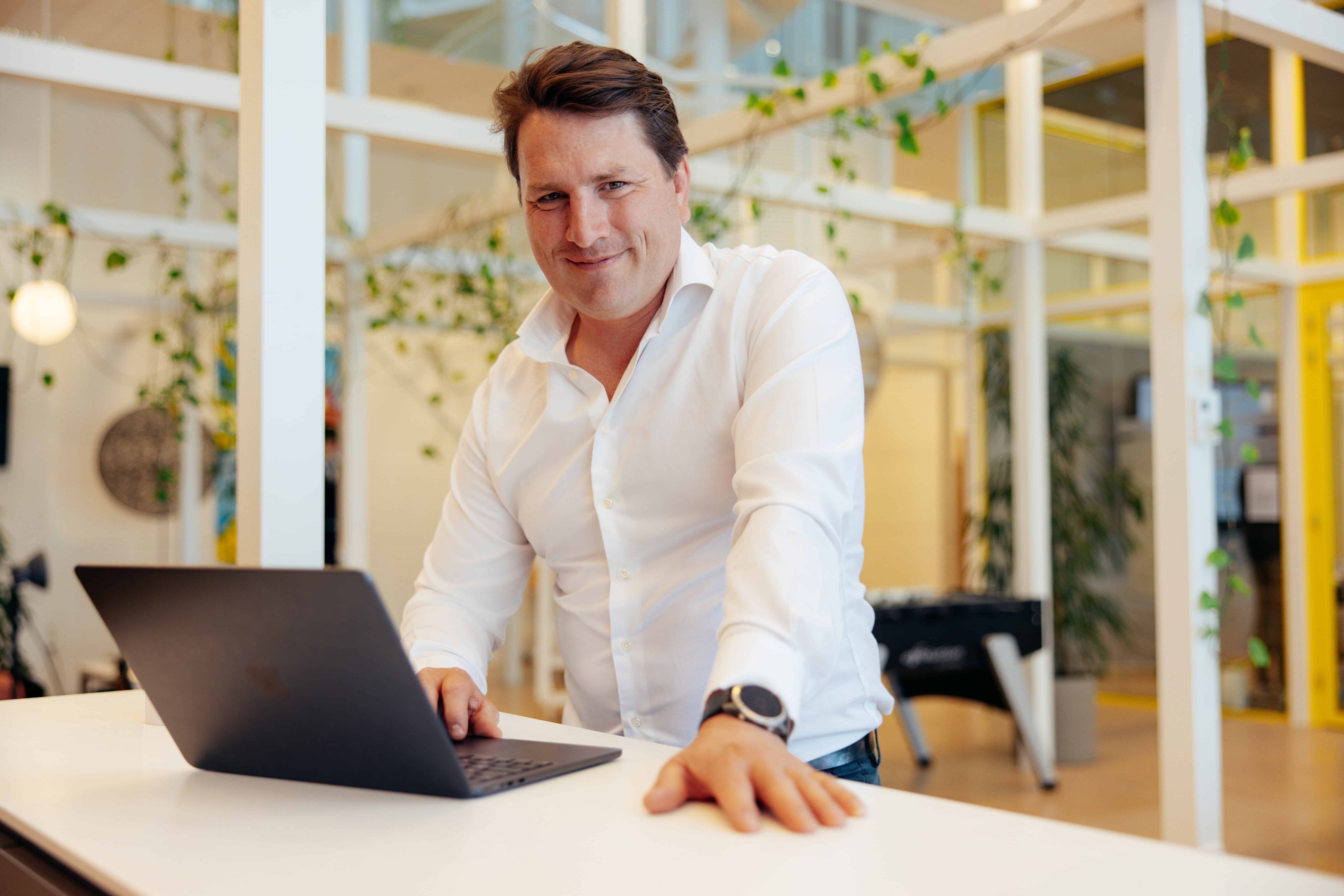 Man in wit overhemd staat achter zijn laptop en steekt zijn linkerarm op tafel.